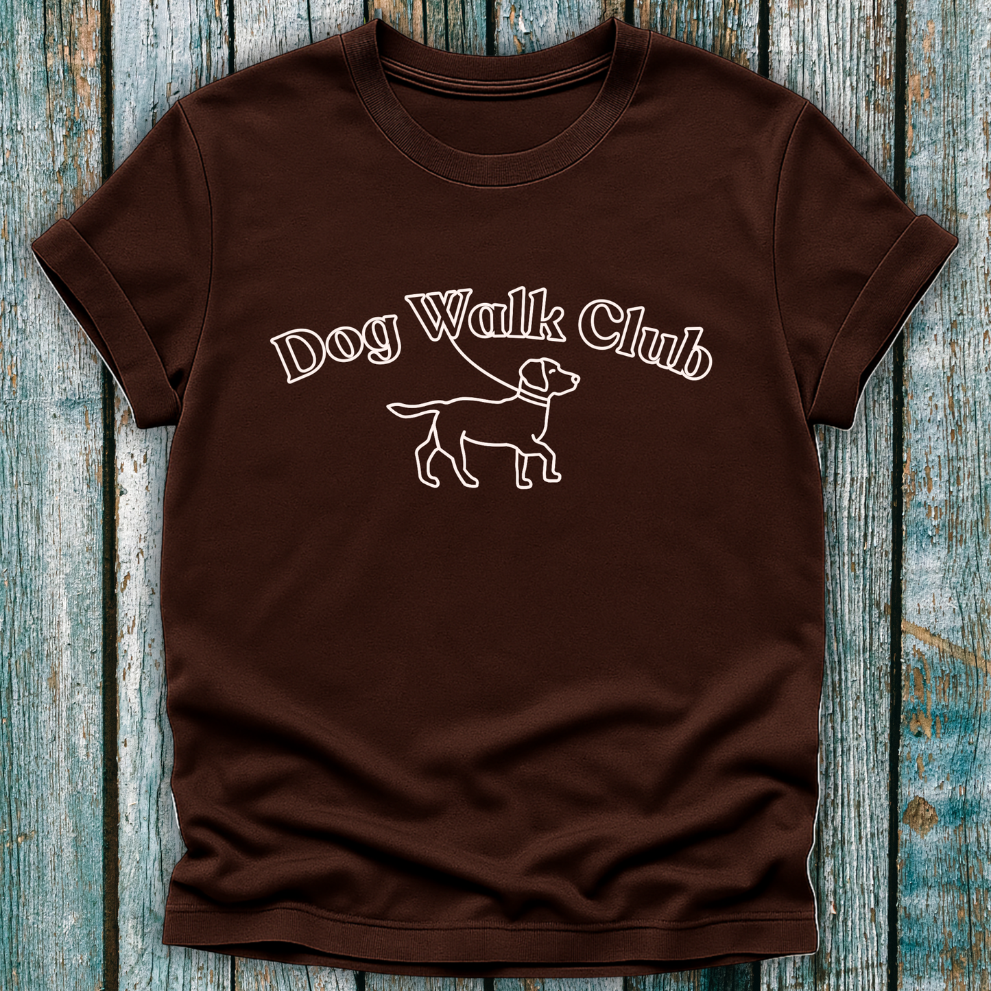 Brown t-shirt with 'Dog Walk Club' text and dog illustration on a wooden surface