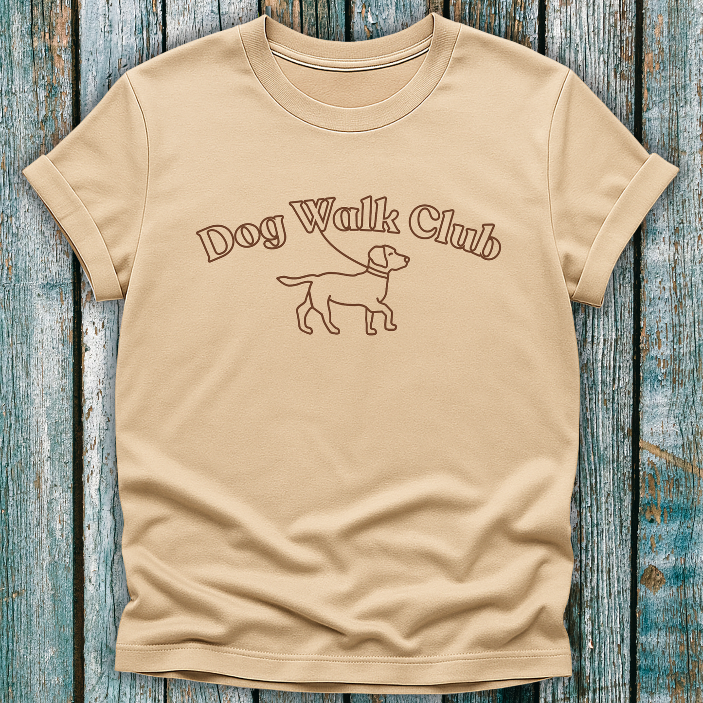 Beige t-shirt with 'Dog Walk Club' text and dog illustration on a wooden surface