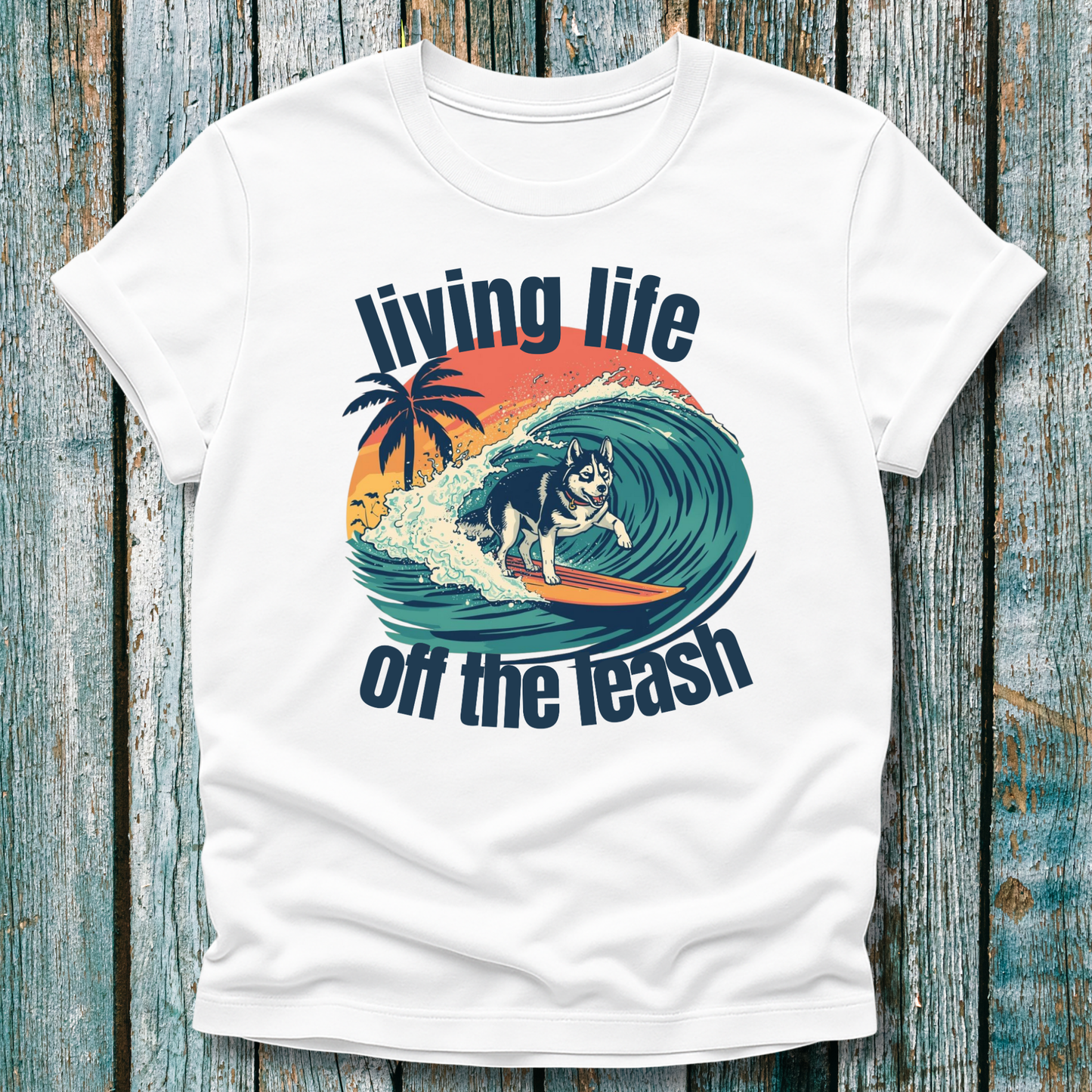 White t-shirt with a graphic of a dog surfing and text 'living life off the leash' on a wooden background