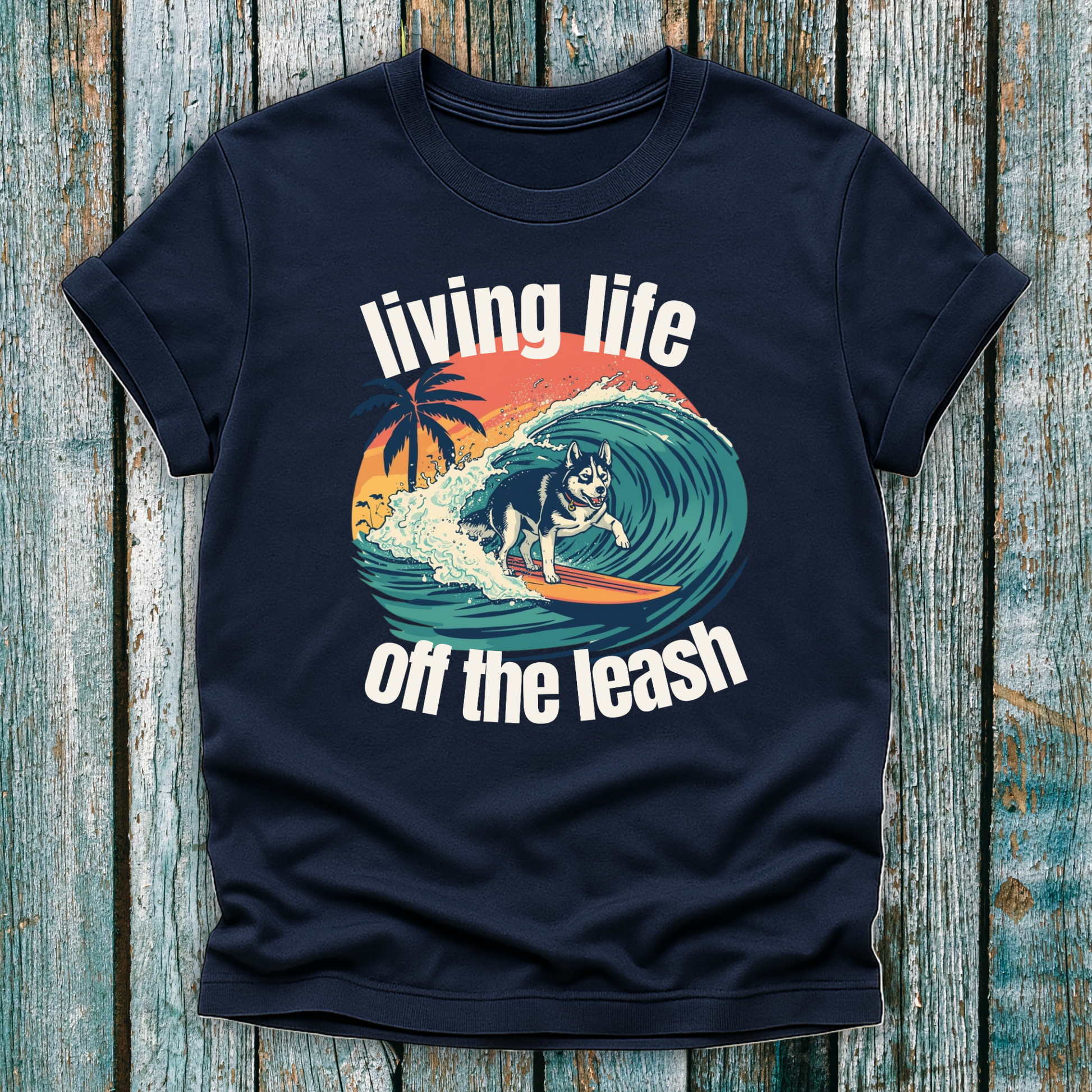 Navy blue t-shirt with a graphic of a dog surfing and text 'living life off the leash' on a wooden surface.