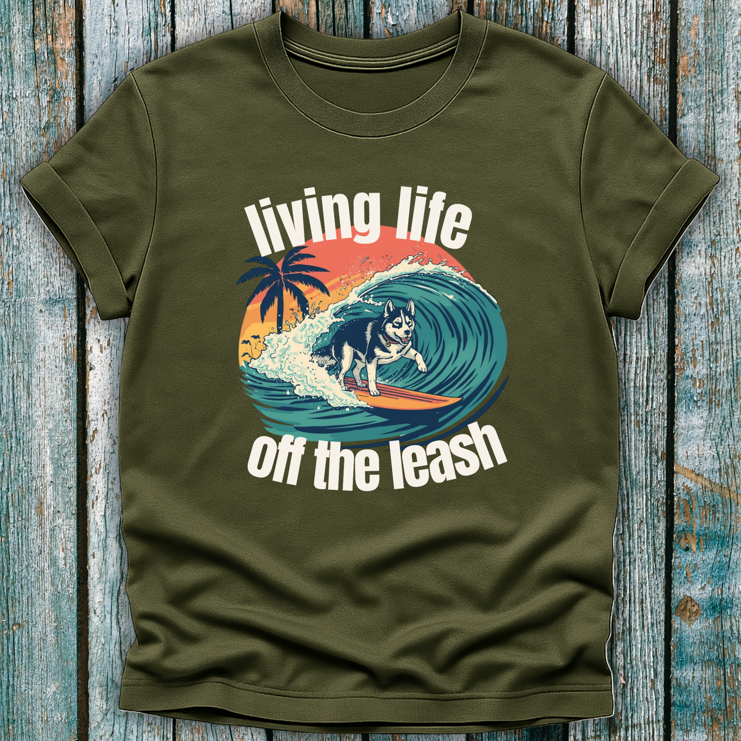 Green t-shirt with a graphic of a dog surfing and text 'living life off the leash' on a wooden surface.