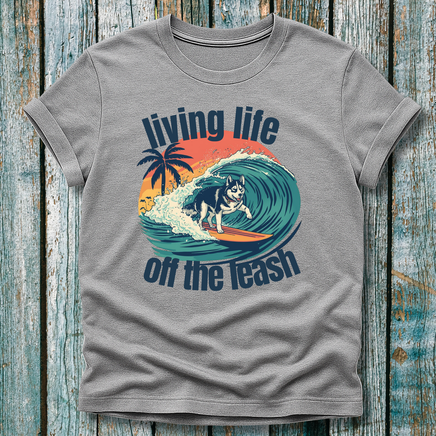 Gray t-shirt with a graphic of a dog surfing and text 'living life off the leash' on a wooden background
