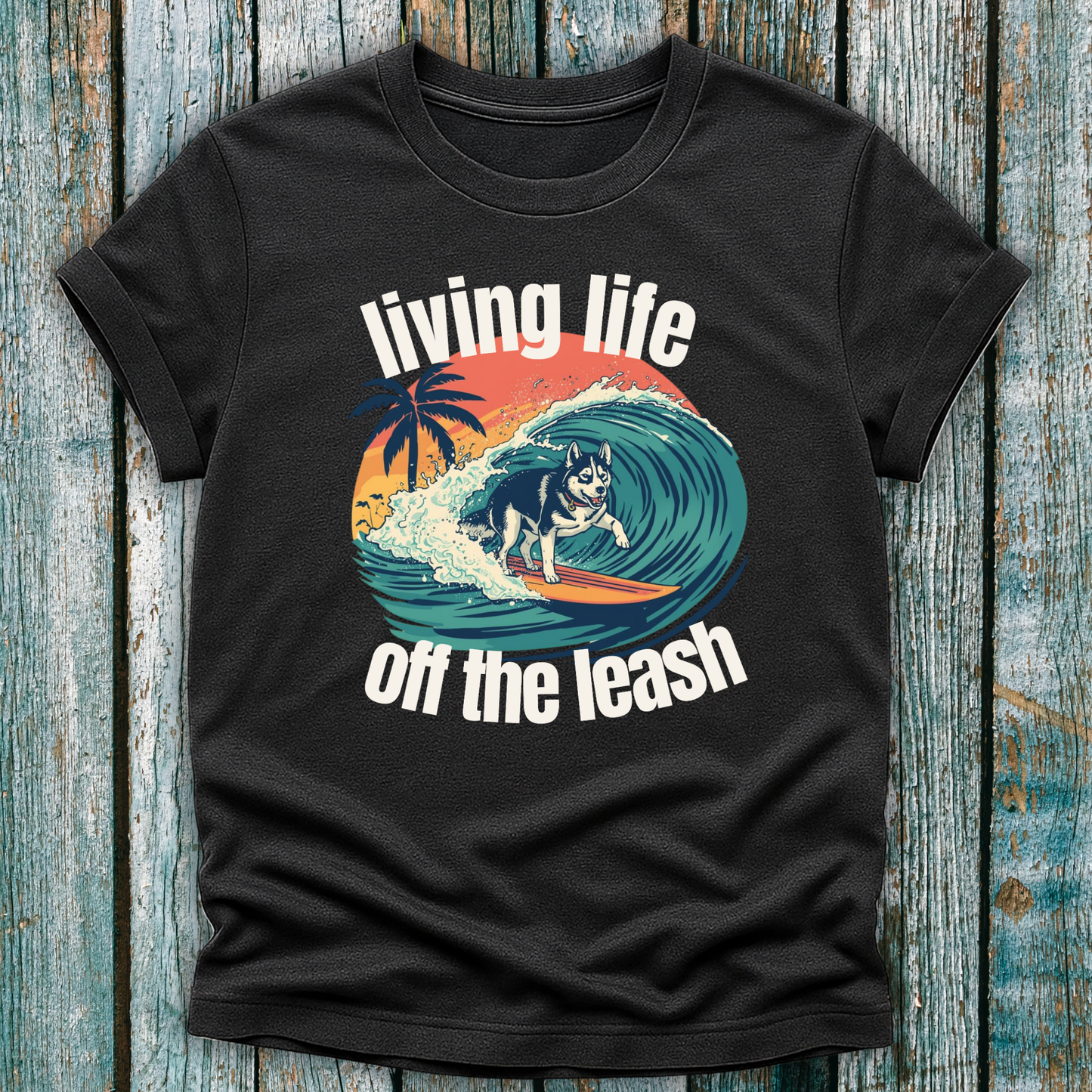 Dark grey t-shirt with a graphic of a dog surfing and text 'living life off the leash' on a wooden surface.