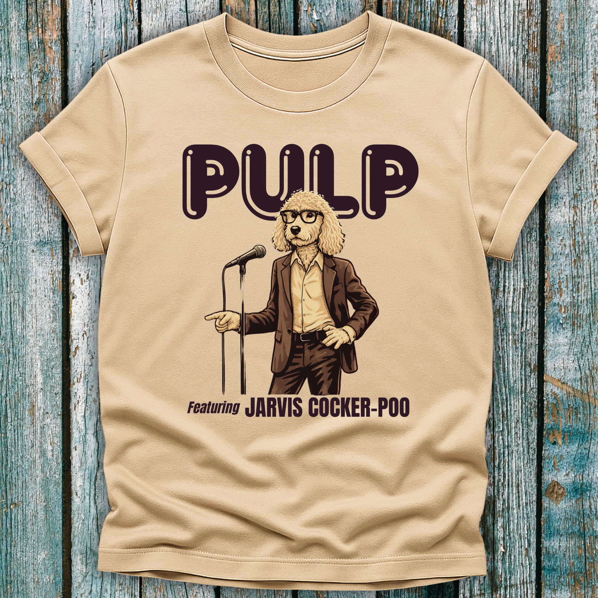 Beige t-shirt with a graphic of a dog in a suit and microphone, labeled 'Pulp featuring Jarvis Cocker Poo', on a wooden surface.