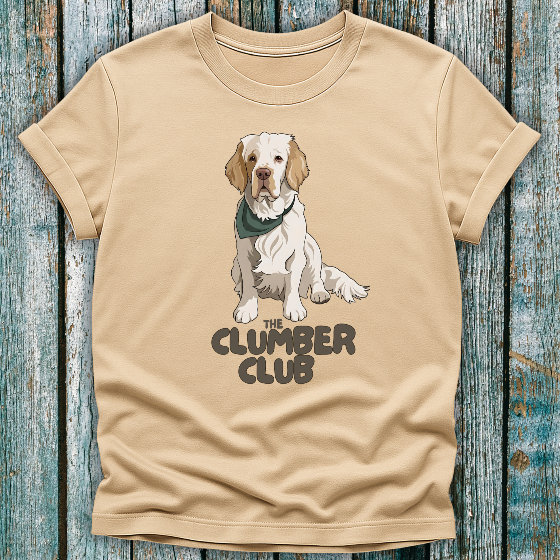 Beige t-shirt with a Clumber Spaniel graphic and 'The Clumber Club' text on a wooden surface