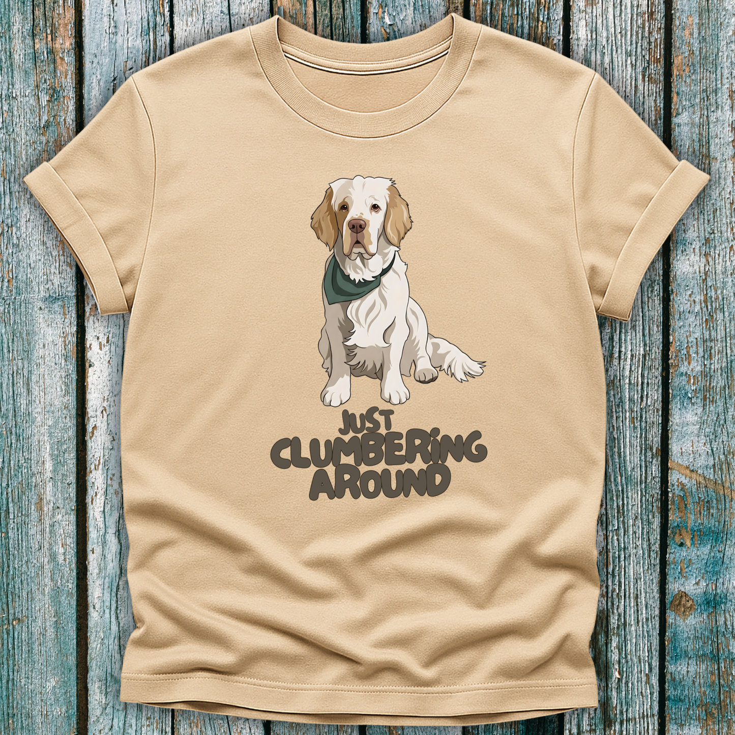 Beige t-shirt with a Clumber Spaniel graphic and text 'Just Clumbering Around' on a wooden surface
