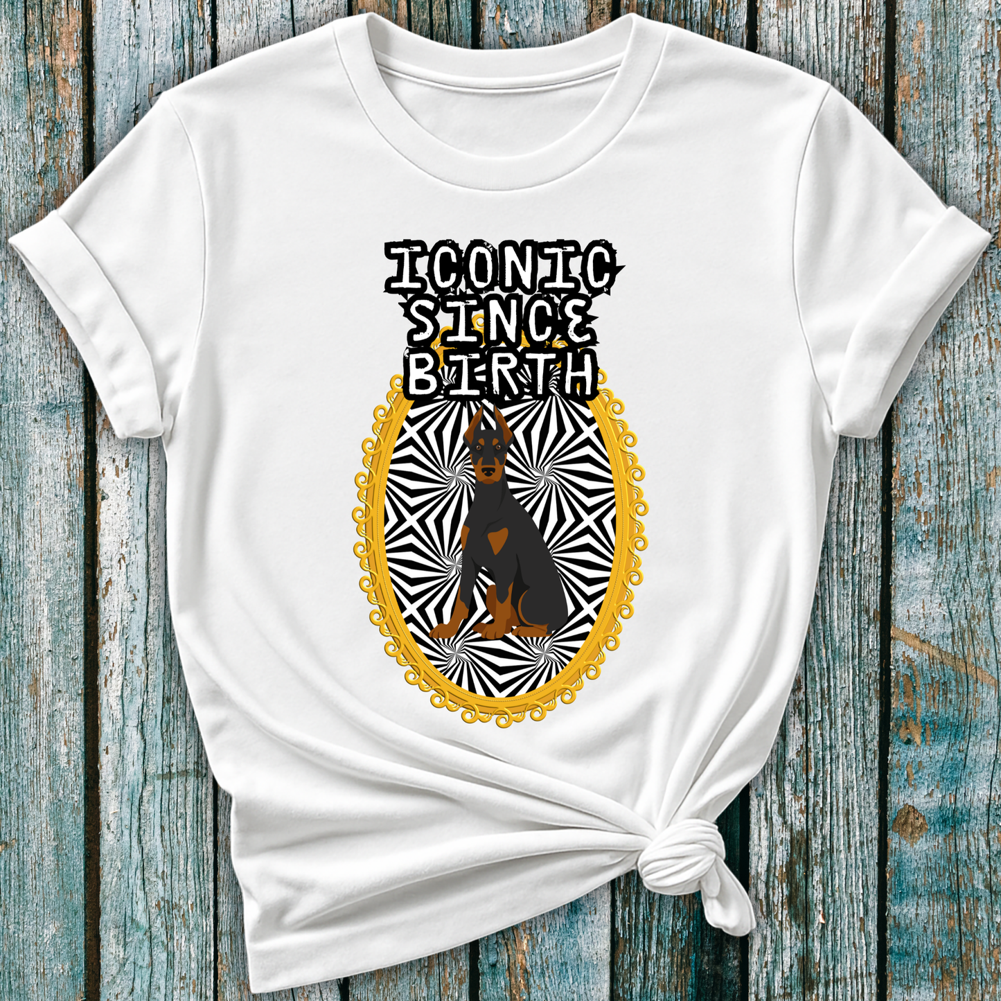 White t-shirt with a graphic design of a Doberman dog and "Iconic Since Birth" text on a wooden surface