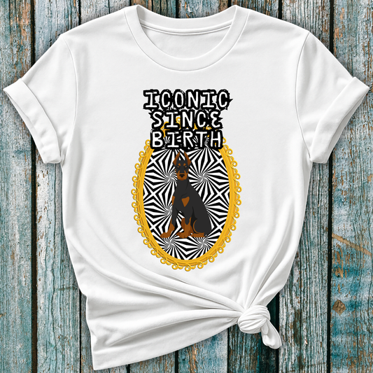 White t-shirt with a graphic design of a Doberman dog and "Iconic Since Birth" text on a wooden surface