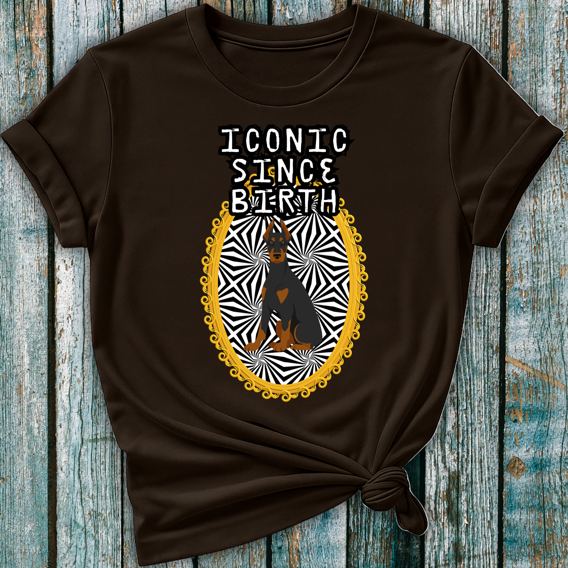 Brown t-shirt with a graphic design of a Doberman dog and "Iconic Since Birth" text on a wooden surface