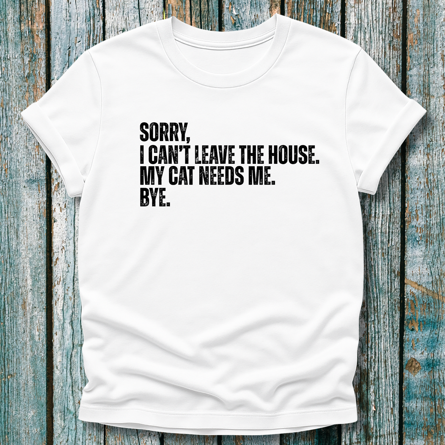 White t-shirt with blacl text "Sorry' I can't leave the house. My cat needs me. Bye." on a wooden background
