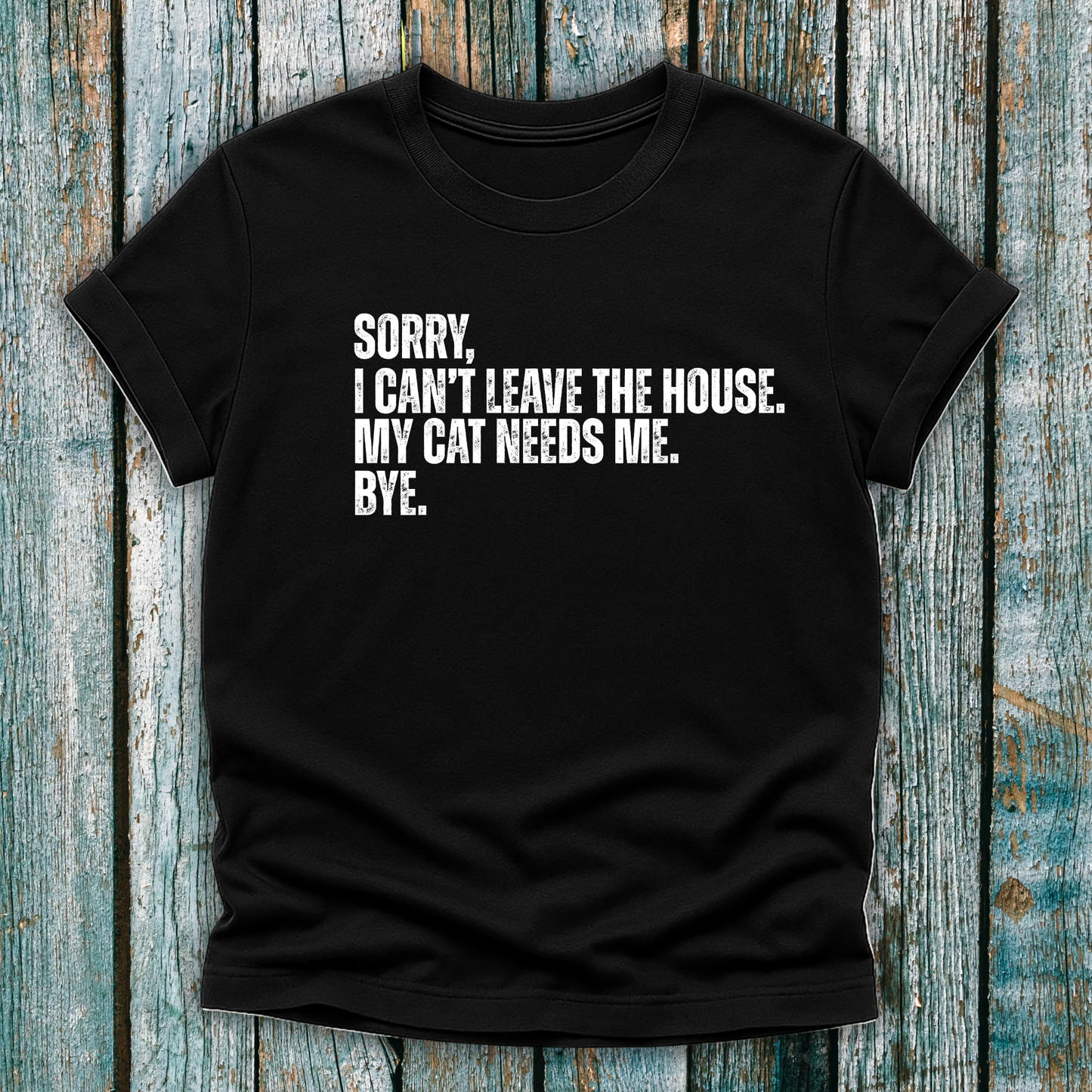 Black t-shirt with white text "Sorry' I can't leave the house. My cat needs me. Bye." on a wooden background