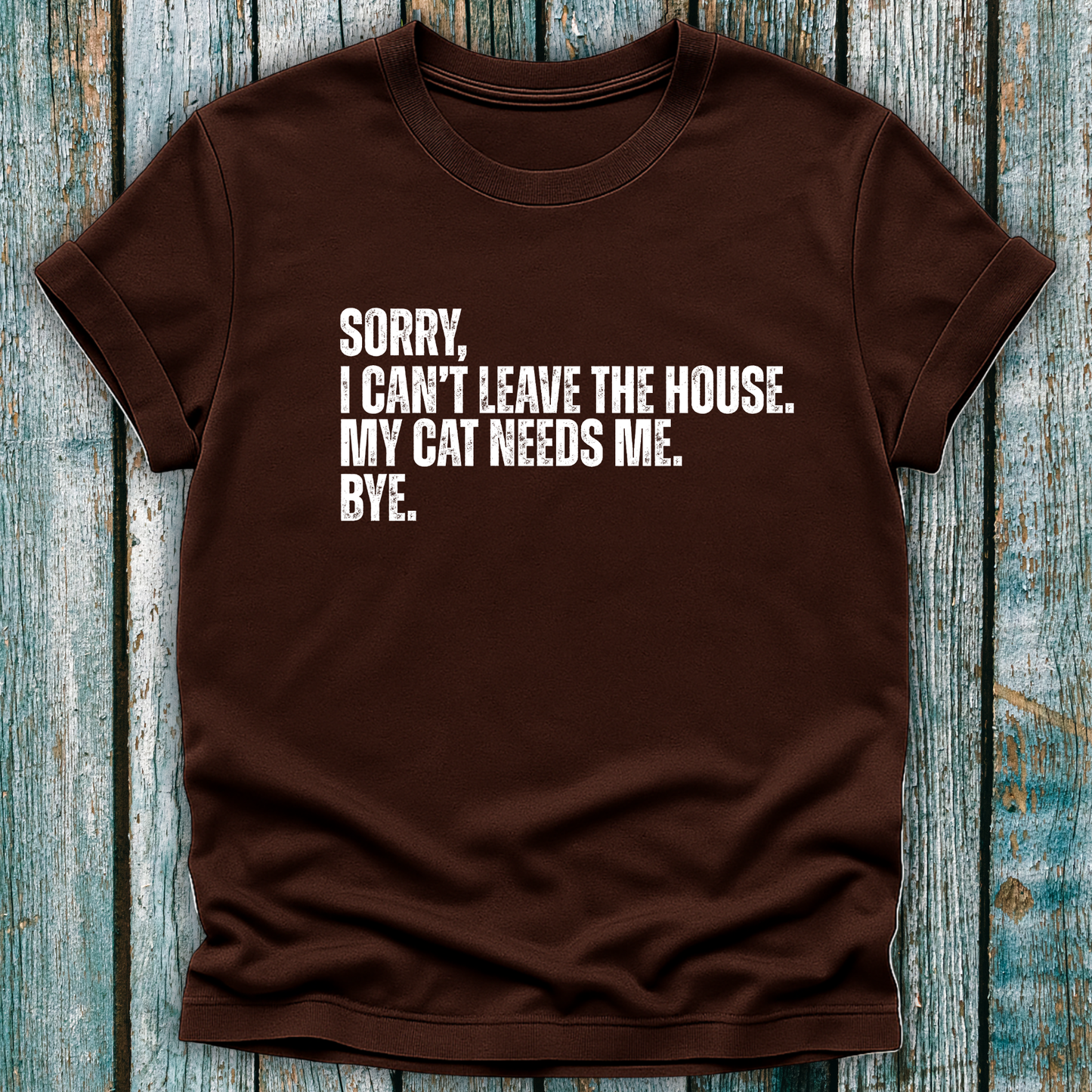 Brown t-shirt with white text "Sorry' I can't leave the house. My cat needs me. Bye." on a wooden background