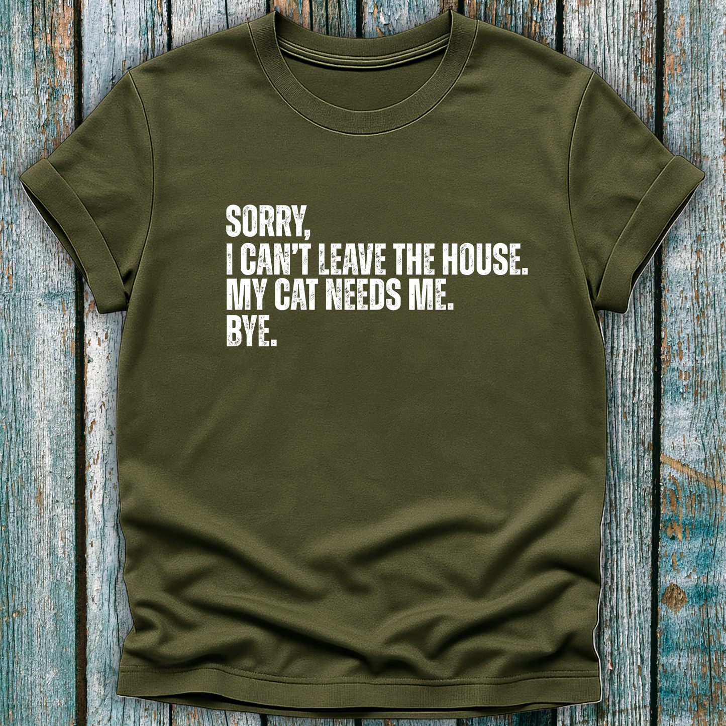 Green t-shirt with white text "Sorry' I can't leave the house. My cat needs me. Bye." on a wooden background