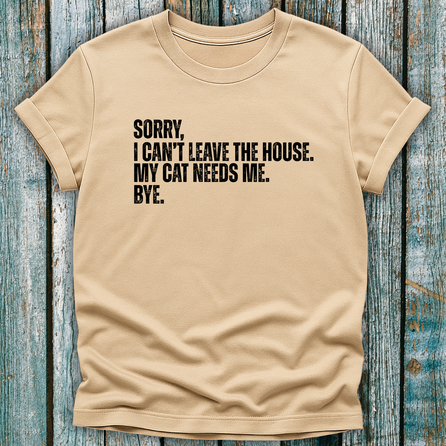 Beige t-shirt with black text "Sorry' I can't leave the house. My cat needs me. Bye." on a wooden background
