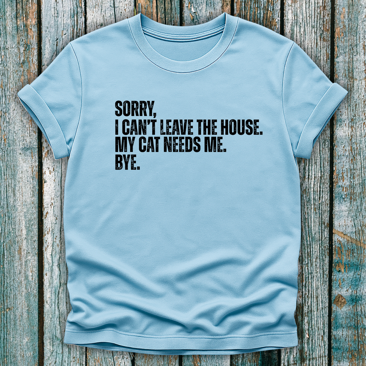 Light blue t-shirt with white text "Sorry' I can't leave the house. My cat needs me. Bye." on a wooden background