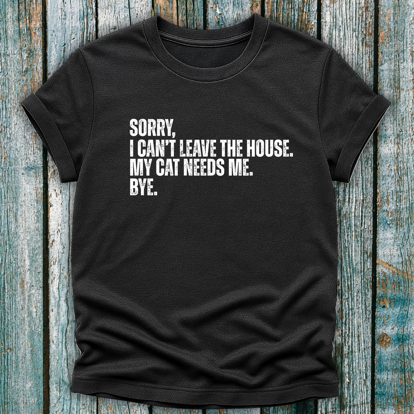 Dark gray t-shirt with white text "Sorry' I can't leave the house. My cat needs me. Bye." on a wooden background