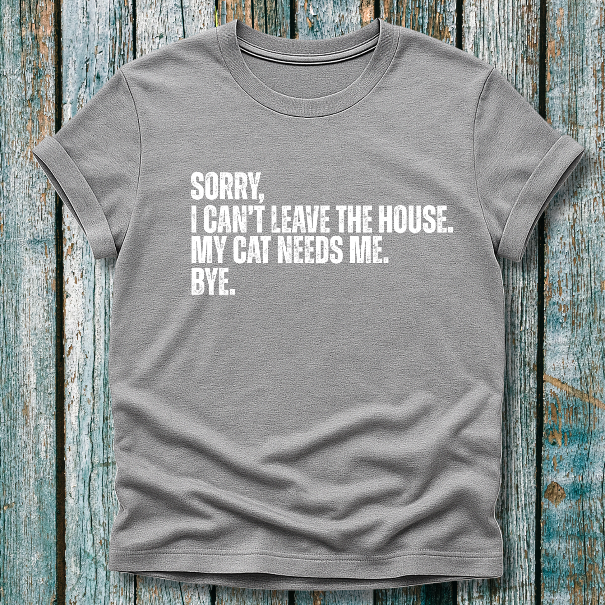 Gray t-shirt with white text "Sorry' I can't leave the house. My cat needs me. Bye." on a wooden background