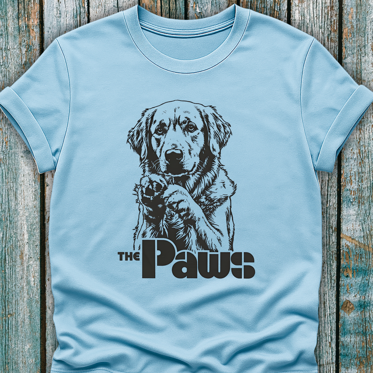 Light blue t-shirt with a dog graphic and 'The Paws' text on a wooden surface