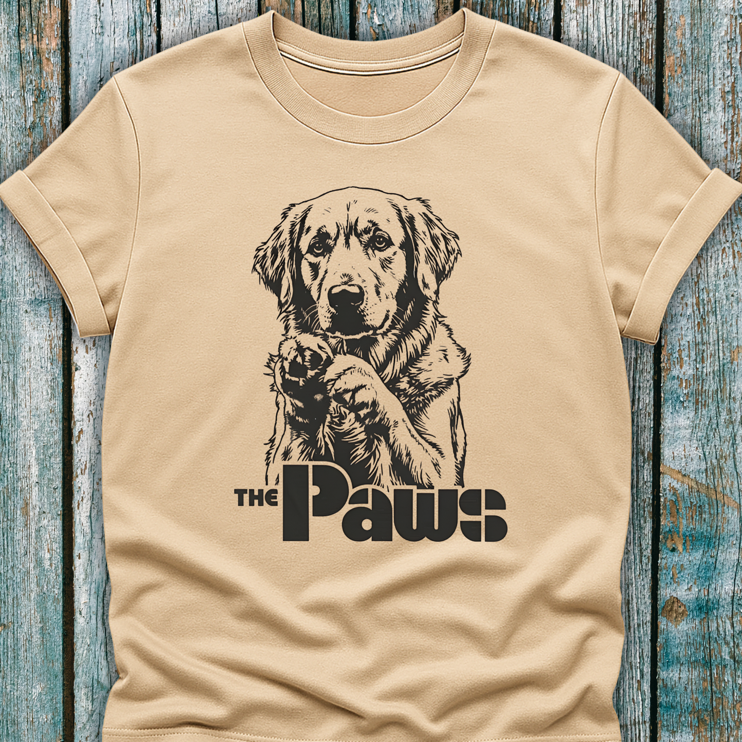 Beige t-shirt with a dog graphic and 'The Paws' text on a wooden surface