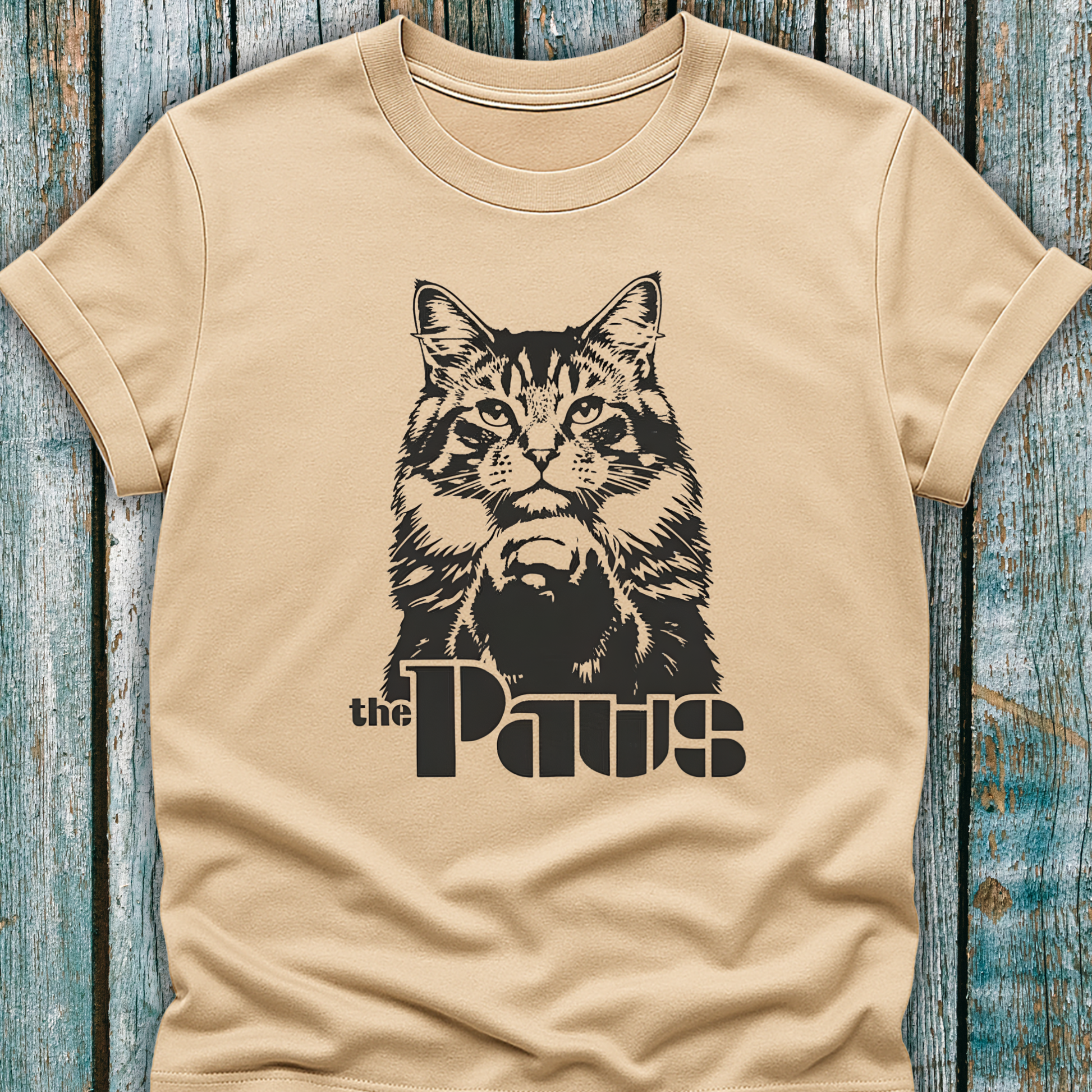Beige t-shirt with a cat graphic and 'the Paws' text on a wooden surface